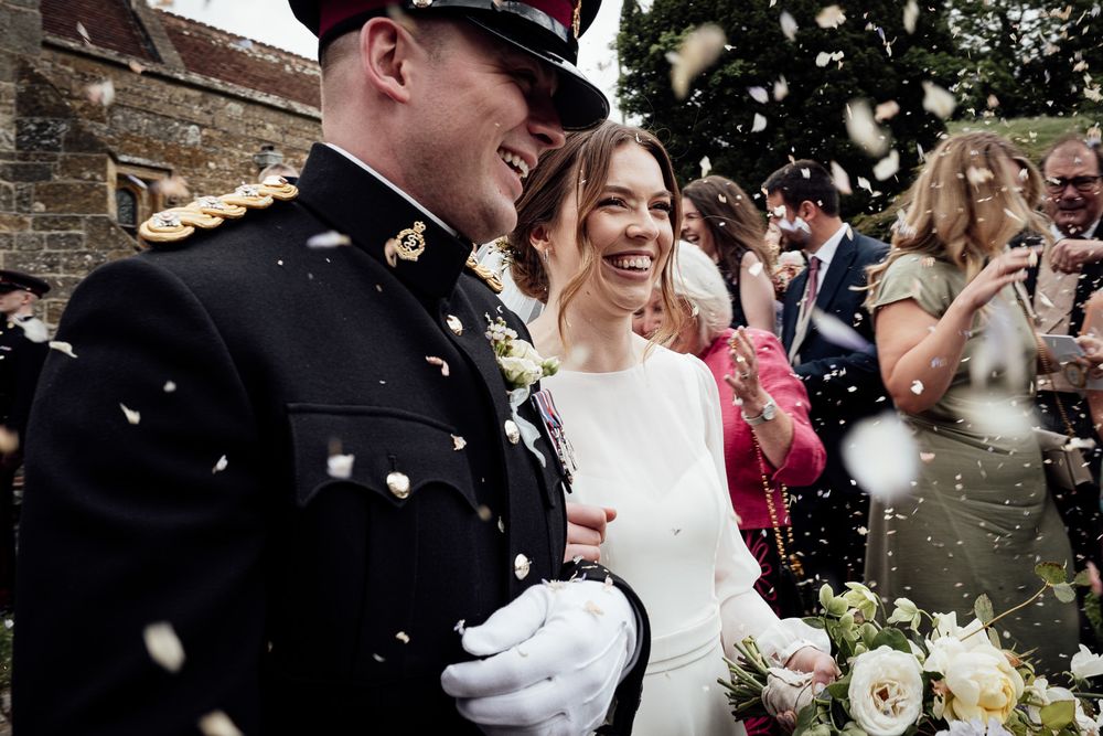 A military officer and bride celebrate their wedding day as guests shower them with confetti outside a brick church.