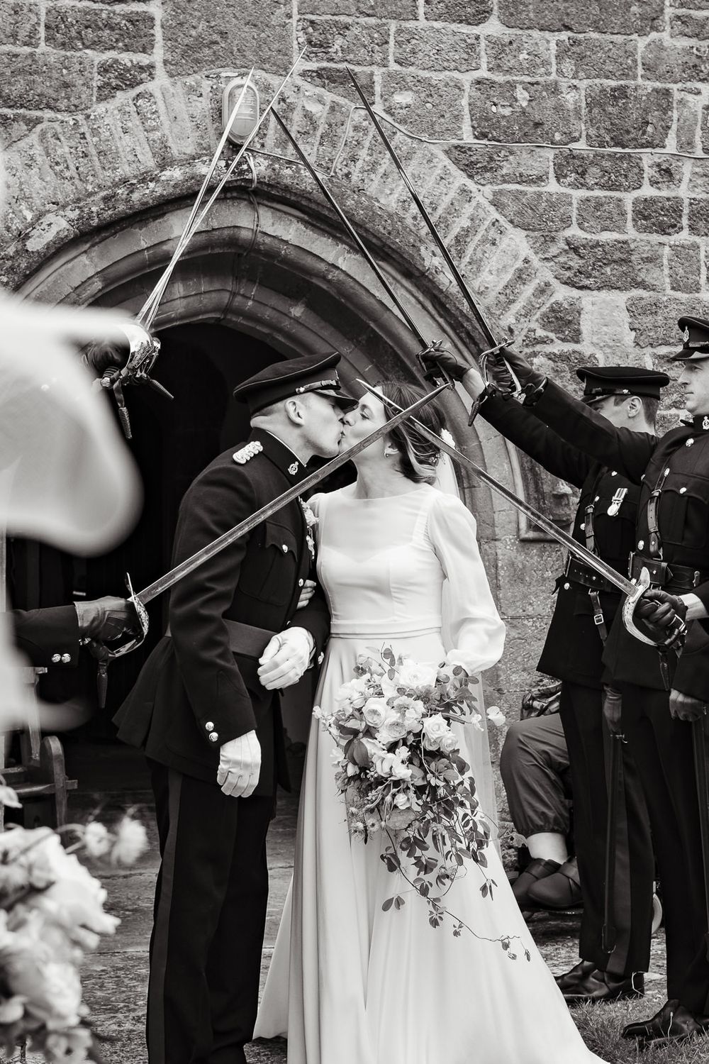 Military officers with swords form an arch for a newlywed couple outside a stone church after their wedding ceremony.