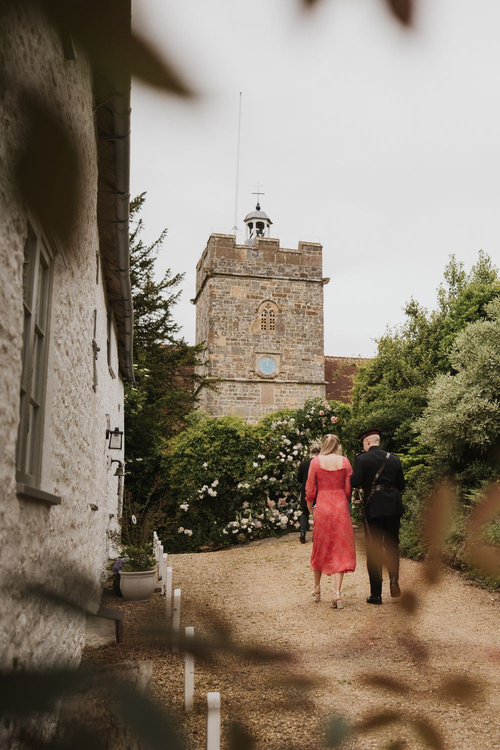 A couple walks along a rustic stone path leading to an ancient stone church tower on a sunny day.