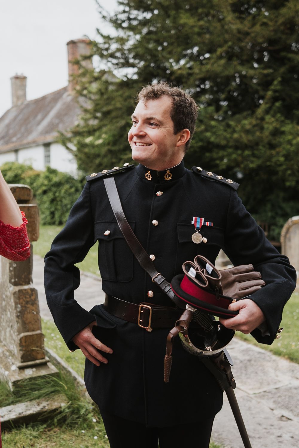 Military service member in dress uniform holding bagpipes stands smiling outdoors near a traditional cottage.
