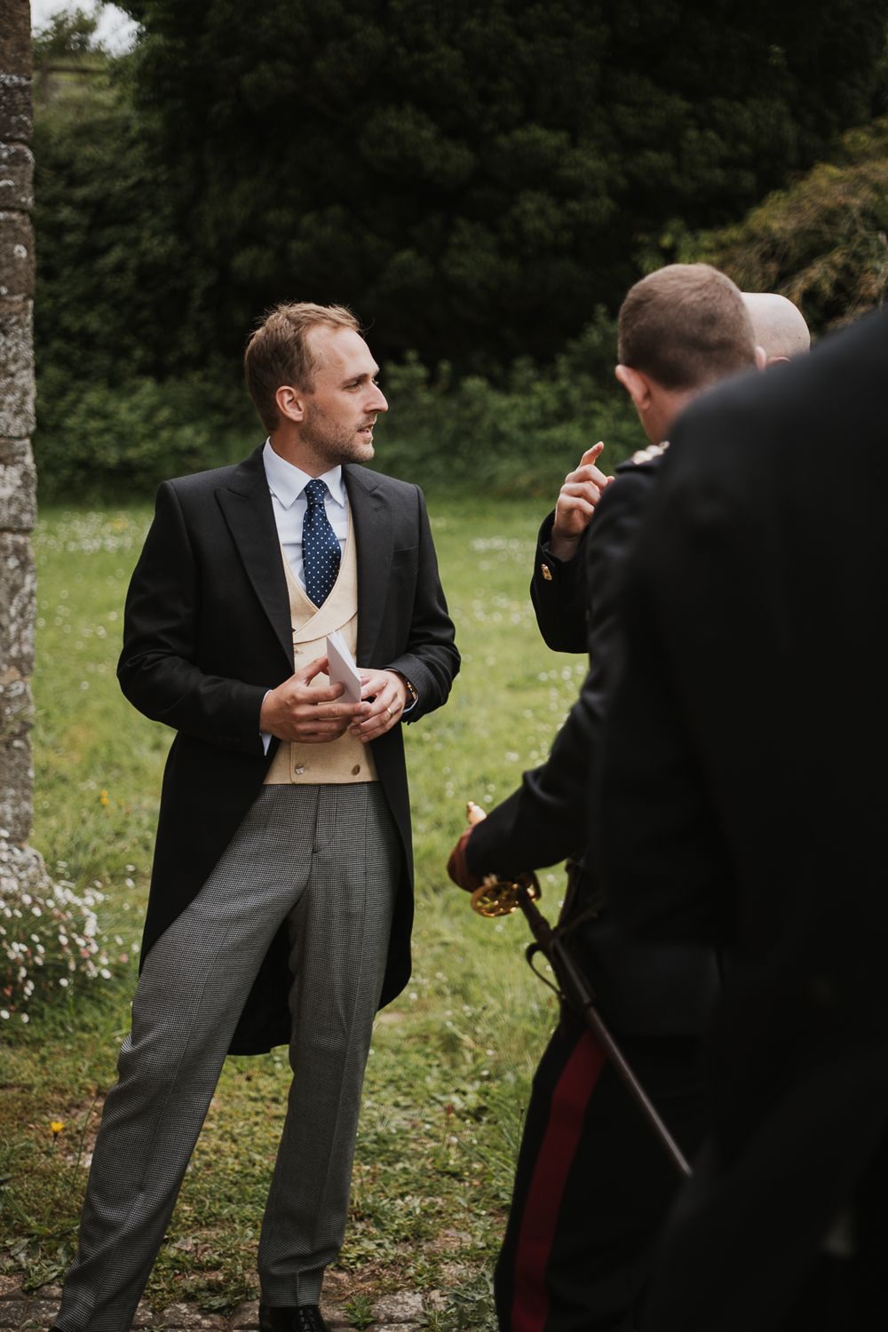 Two men in formal attire engage in a playful sword fight duel outdoors in a garden setting.