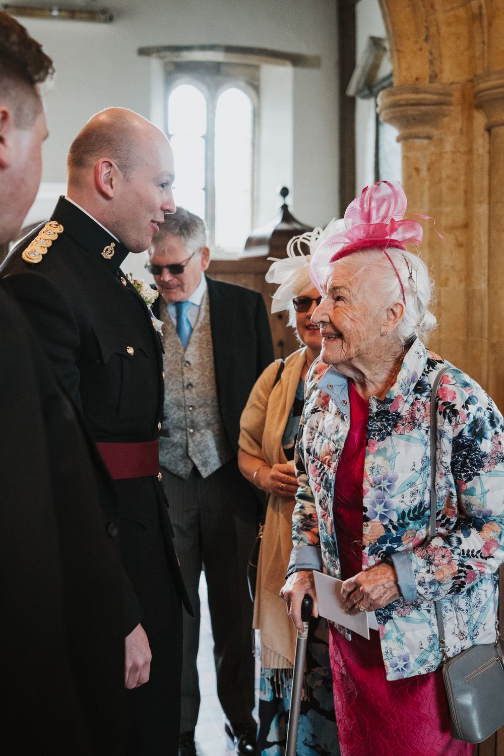A cheerful elderly woman wearing a pink fascinator and floral jacket chats with a uniformed serviceman in a church setting.