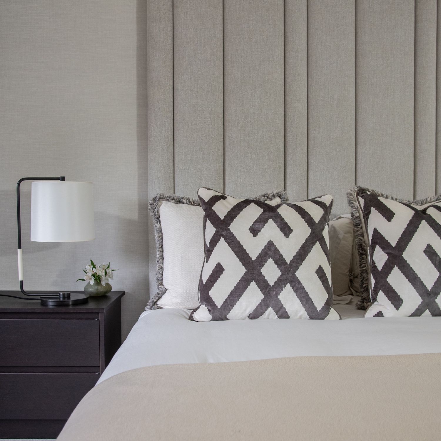 Modern bedroom with geometric black and white patterned throw pillows, white bedding, and a sleek table lamp. Andrew Martin cushions.