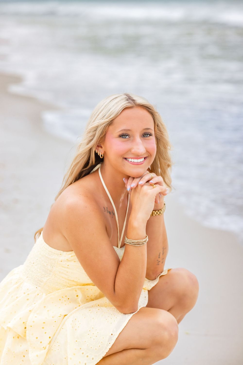A person wearing a light yellow sundress poses casually on a sandy beach with ocean waves in the background.