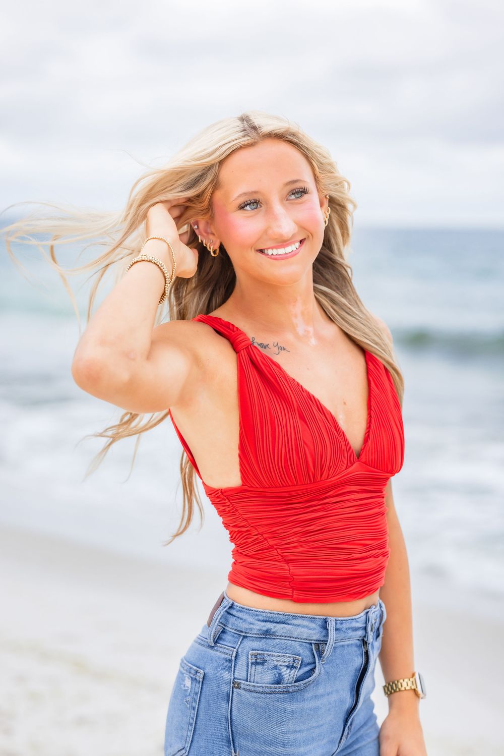 A woman in a red crop top and jeans smiles while standing on a beach with blonde hair blowing in the breeze.