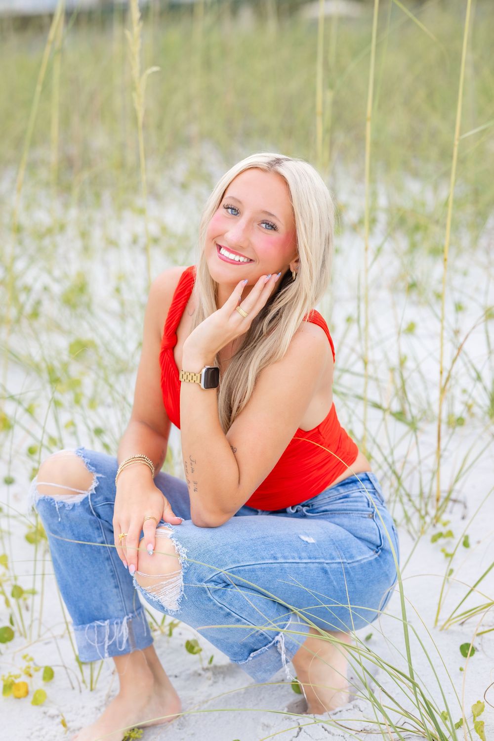 A woman in a red tank top and ripped jeans poses amongst beach grass and sand dunes on a sunny day.