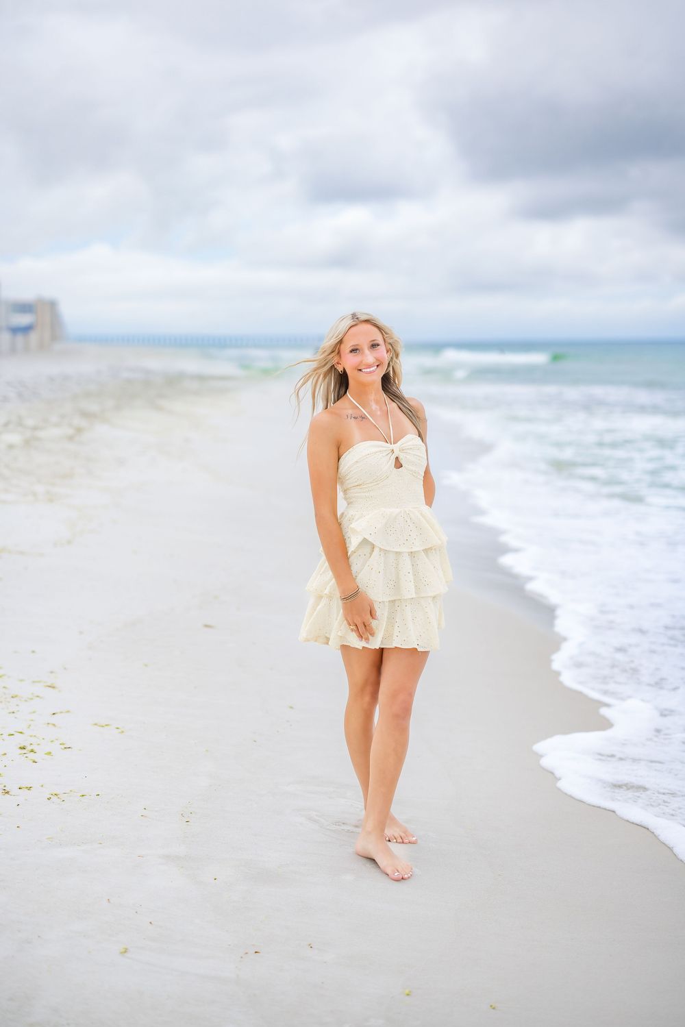 A person in a cream-colored ruffle dress walks barefoot along a cloudy beach shoreline at the ocean's edge.