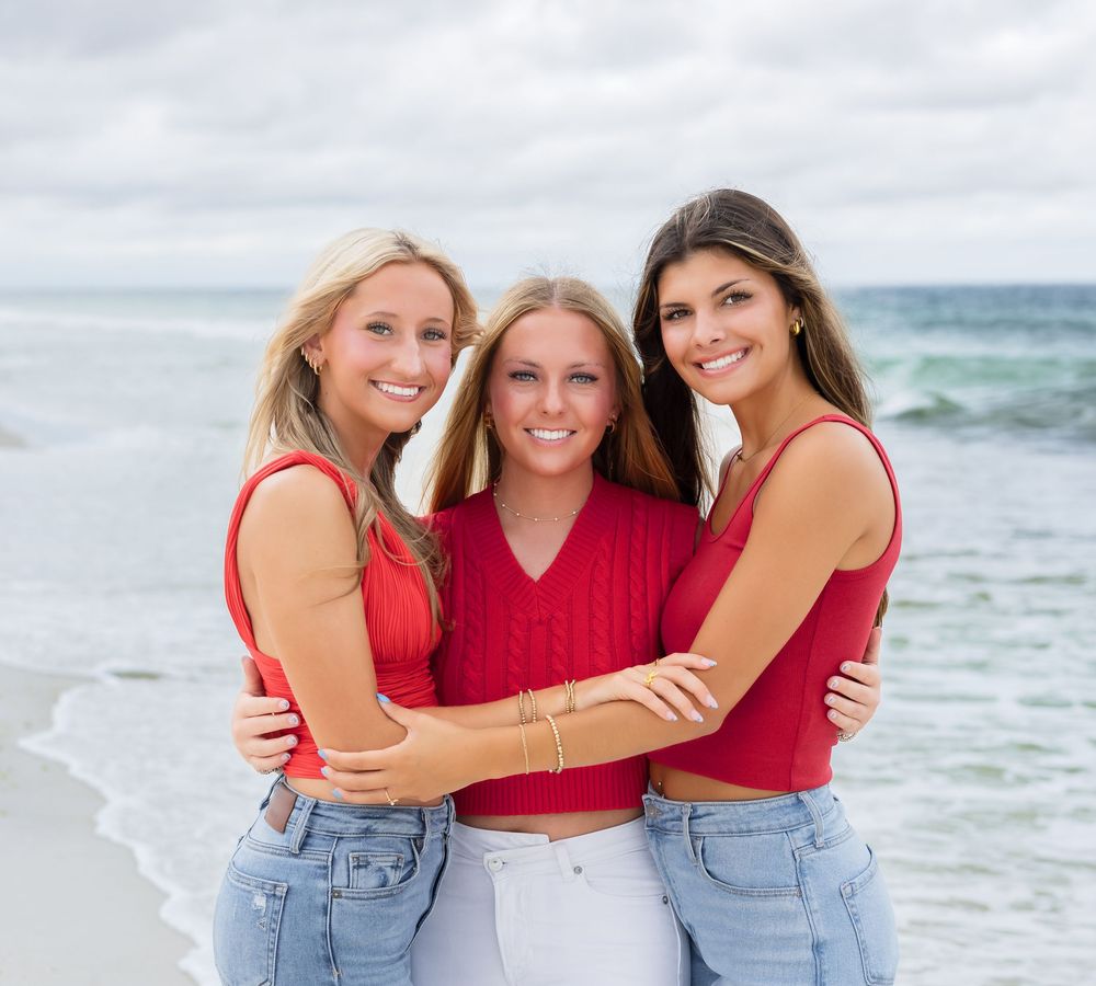 Three people wearing red tops and blue jeans pose together for casual beach portraits at the ocean.