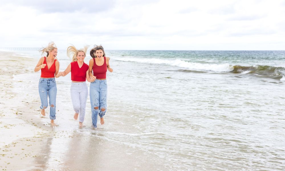 Three friends in red tops and jeans run joyfully along the beach shoreline with ocean waves behind them.