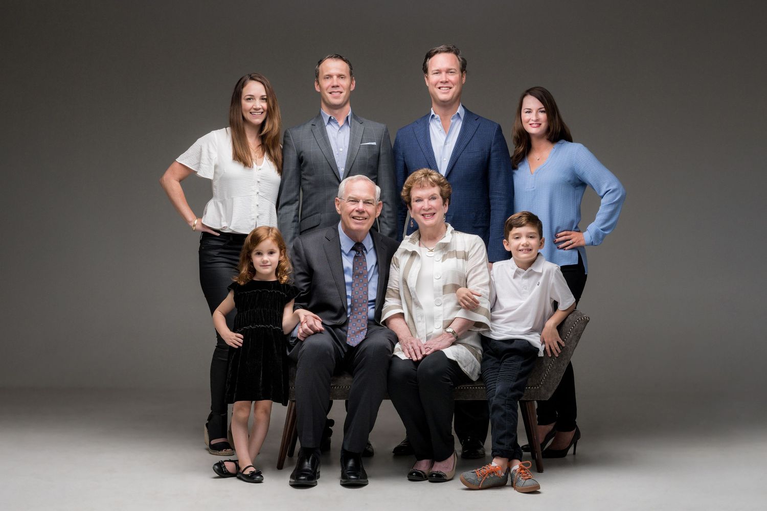 Professionally dressed family members sit together in clean studio portraits against a grey backdrop.