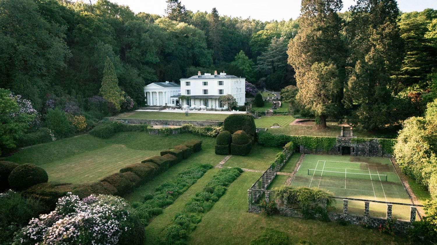 Aerial view of a stately white mansion with manicured gardens and tennis court nestled among lush trees at dusk.