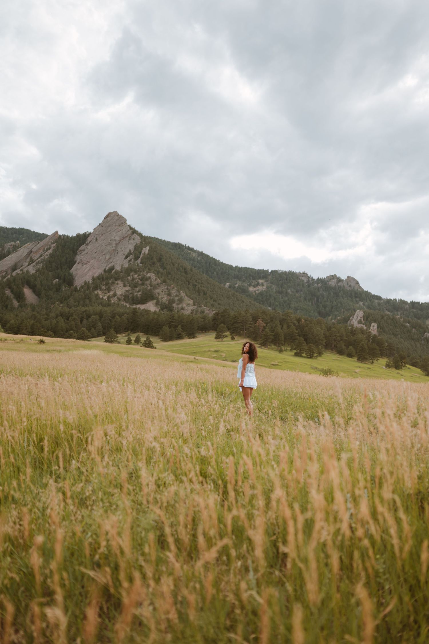 A solitary figure stands in a golden meadow with the dramatic Flatirons rock formation rising in the cloudy Boulder backdrop.