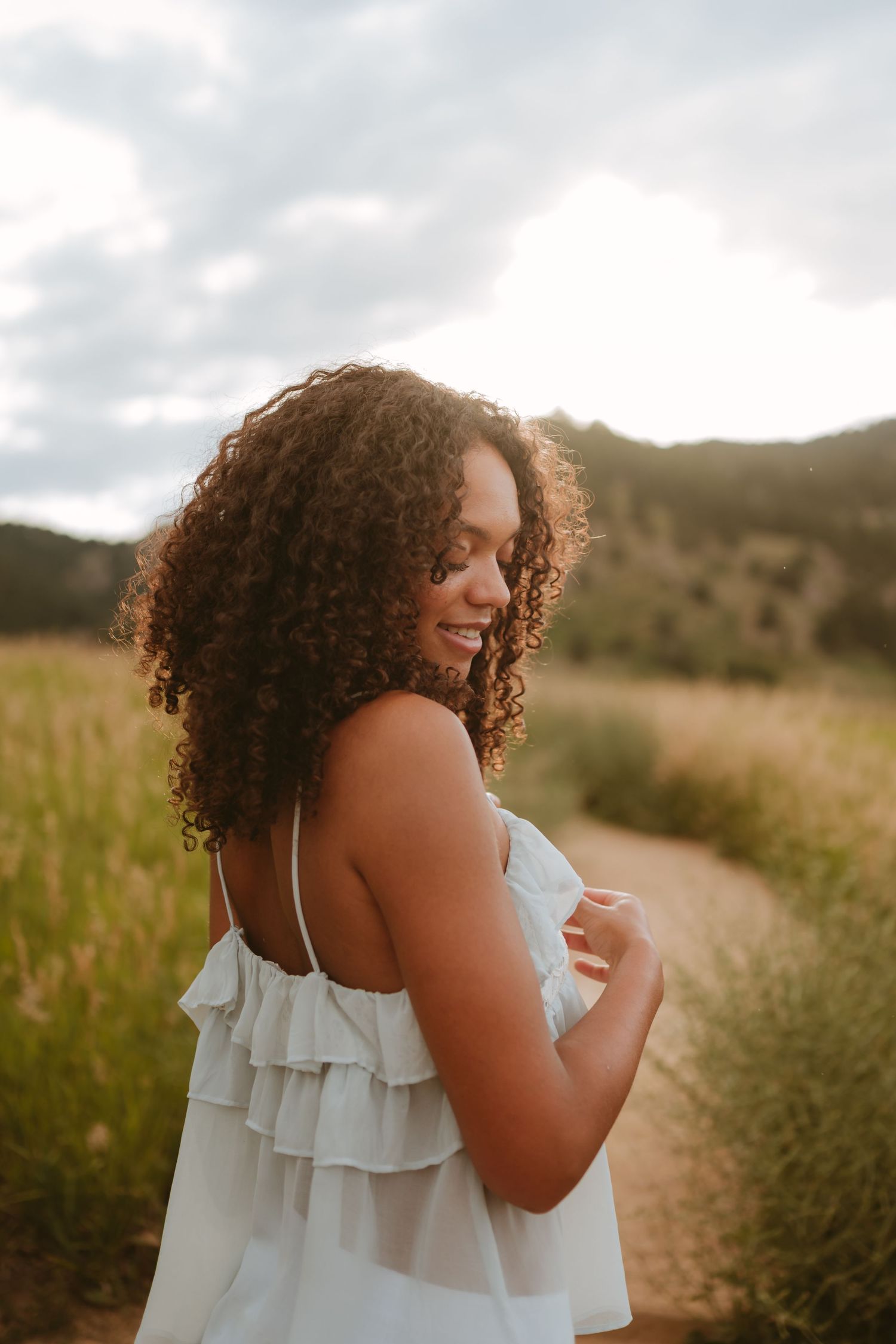 A woman in a white ruffle sundress laughs joyfully while standing in a golden meadow at sunset.
