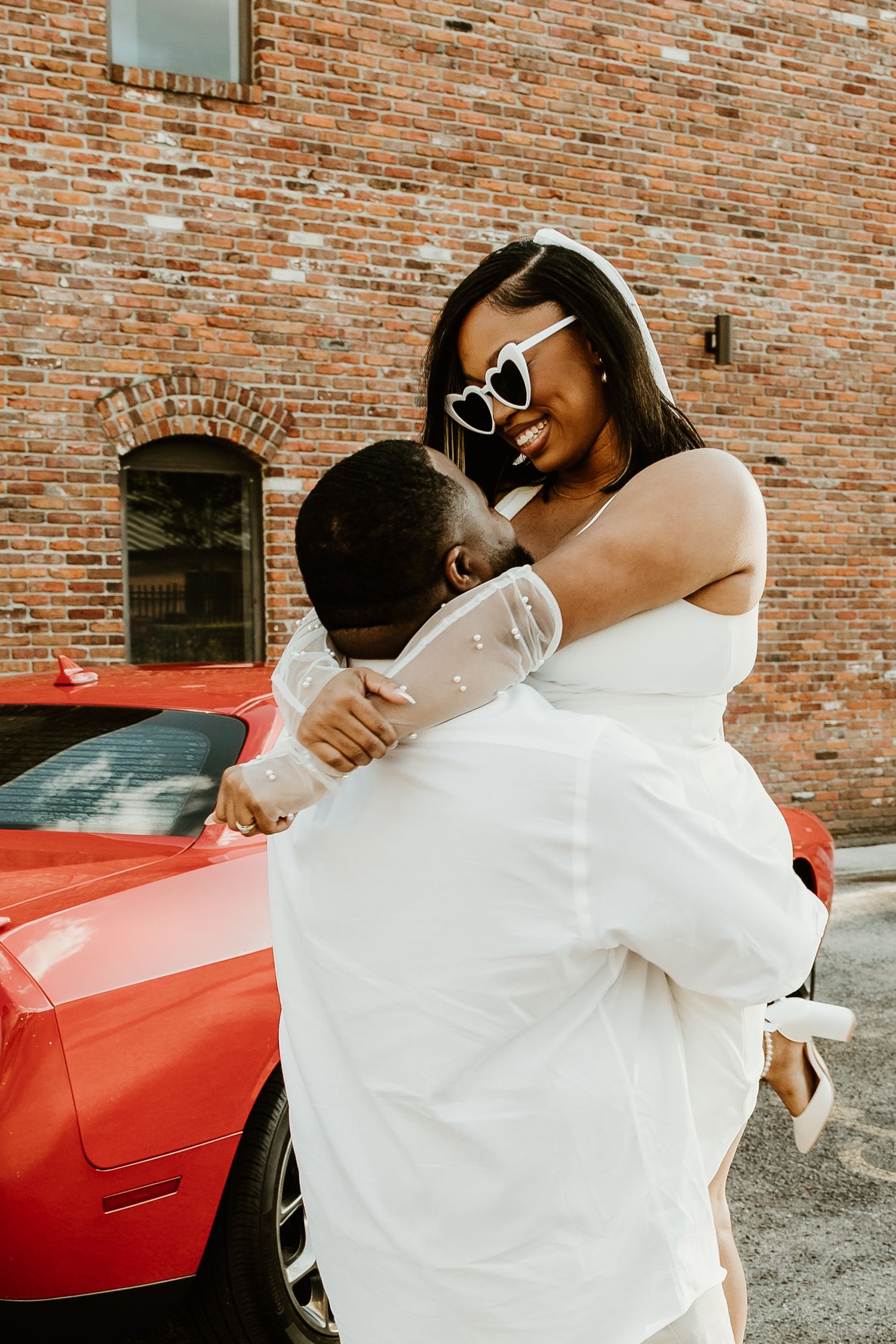 A joyful embrace captured against an exposed brick wall with a red vintage car in the background.