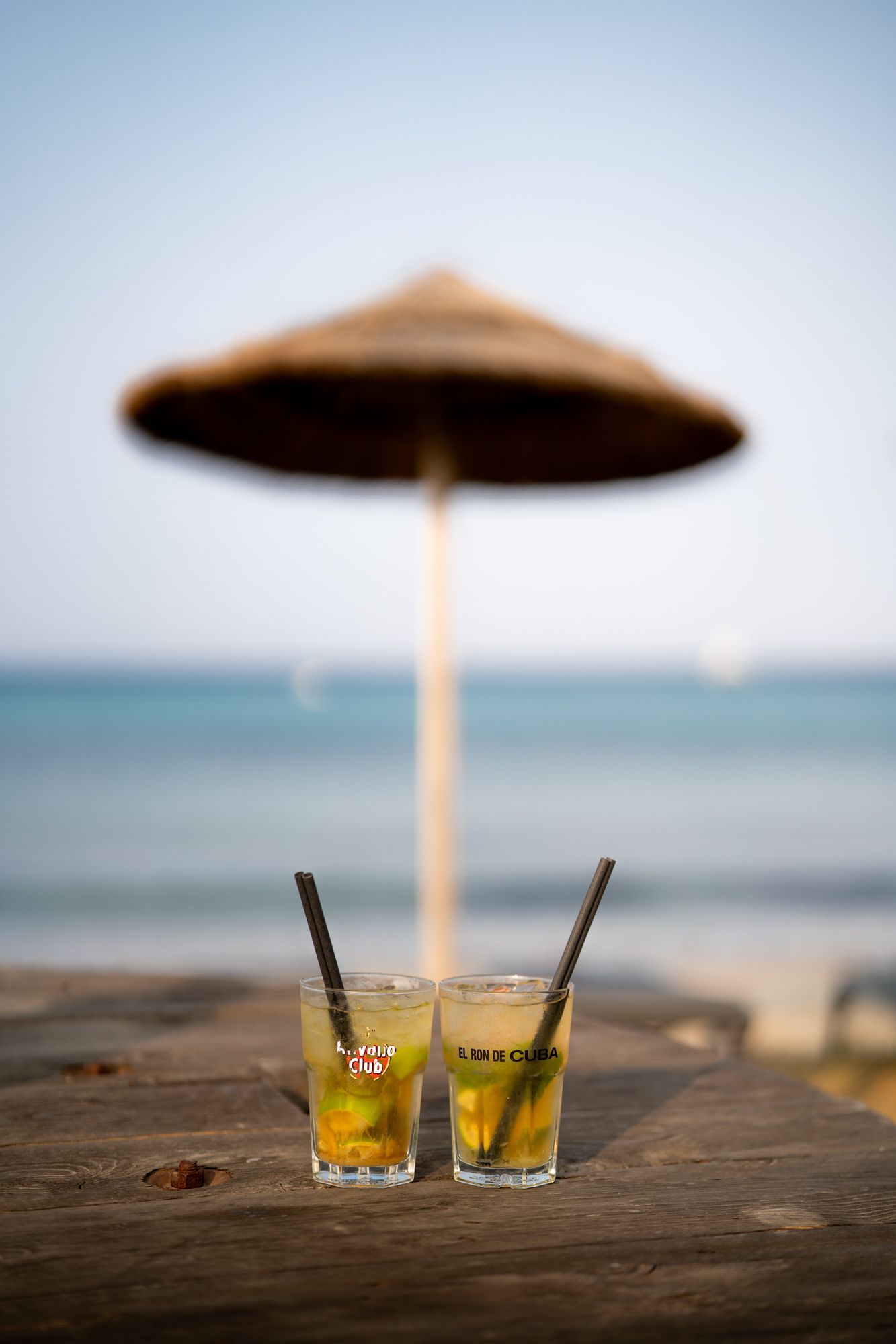 deux mojitos posés sur une table en bois avec en arrière plan un parasol en paille devant la mer turquoise du cap corse