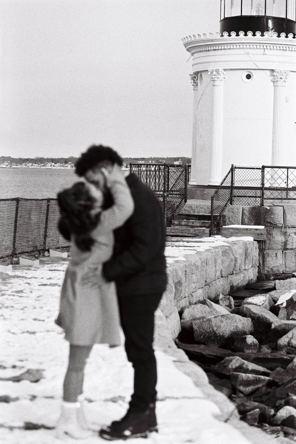 Kissing couple in front of the Bug Light lighthouse in South Portland, Maine. A black and white film photo of their embrace.