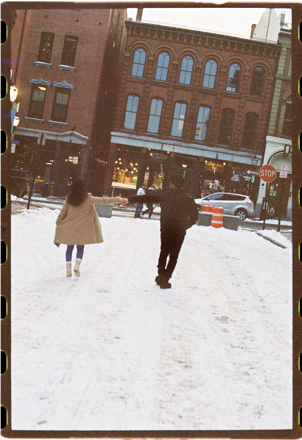 35mm film photograph of a couple walking and playing and holding hands in the snow between two brick architectural buildings in Maine.