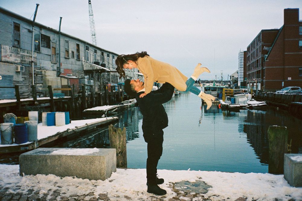 Recently engaged couple poses by holding his fiancé up in the air in the winter on a wharf surrounded by water and fishing boats in Portland