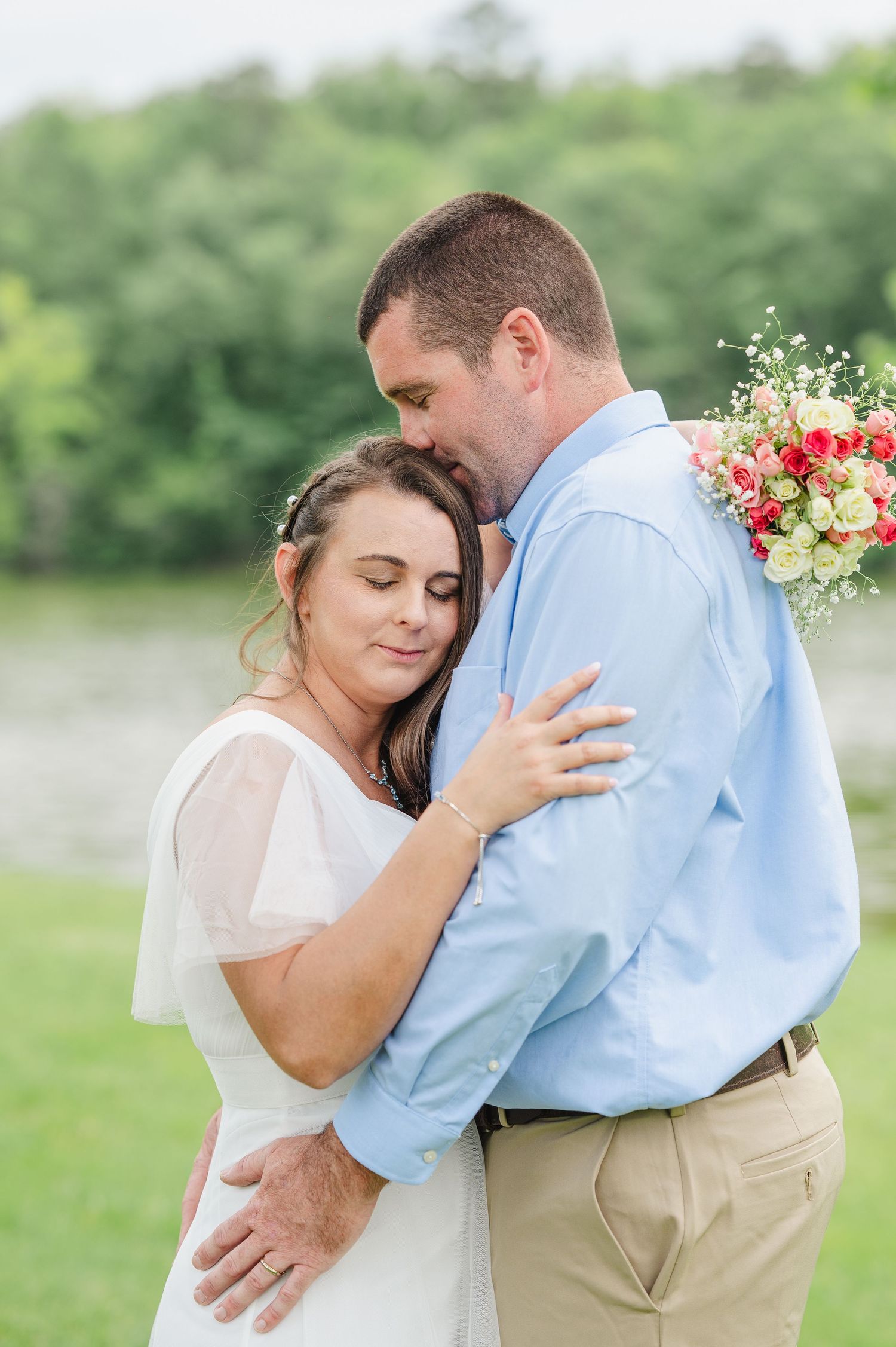 A romantic outdoor moment between a North Carolina couple embracing, with the bride holding a colorful bouquet of flowers by a lake.
