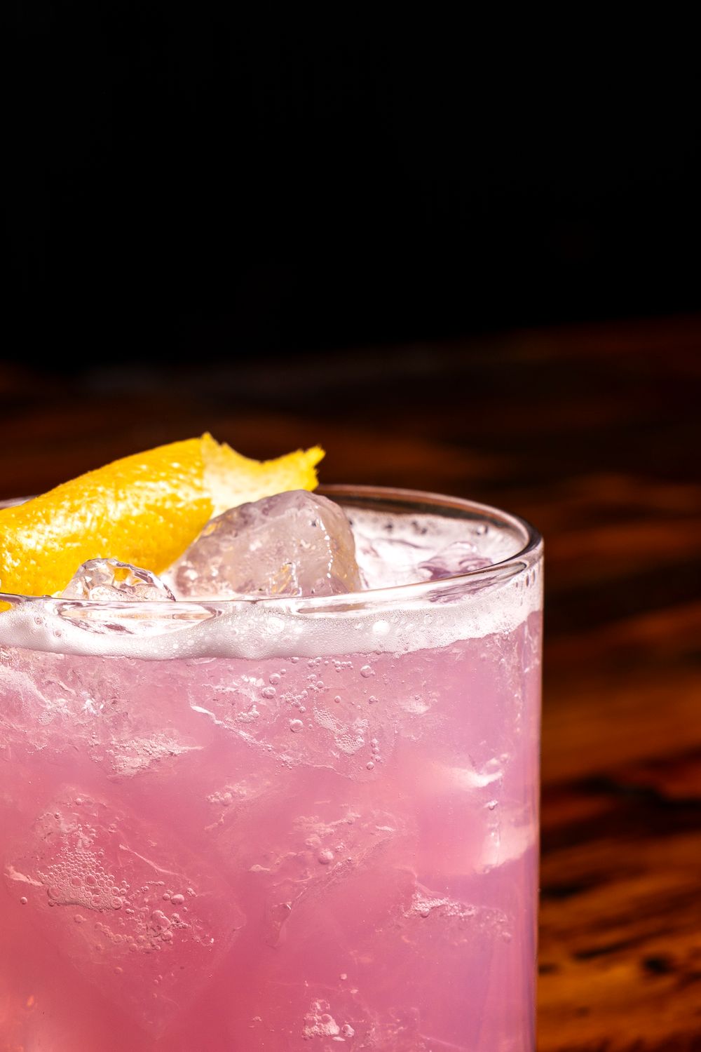 Pink cocktail with ice and orange garnish on dark wooden surface.