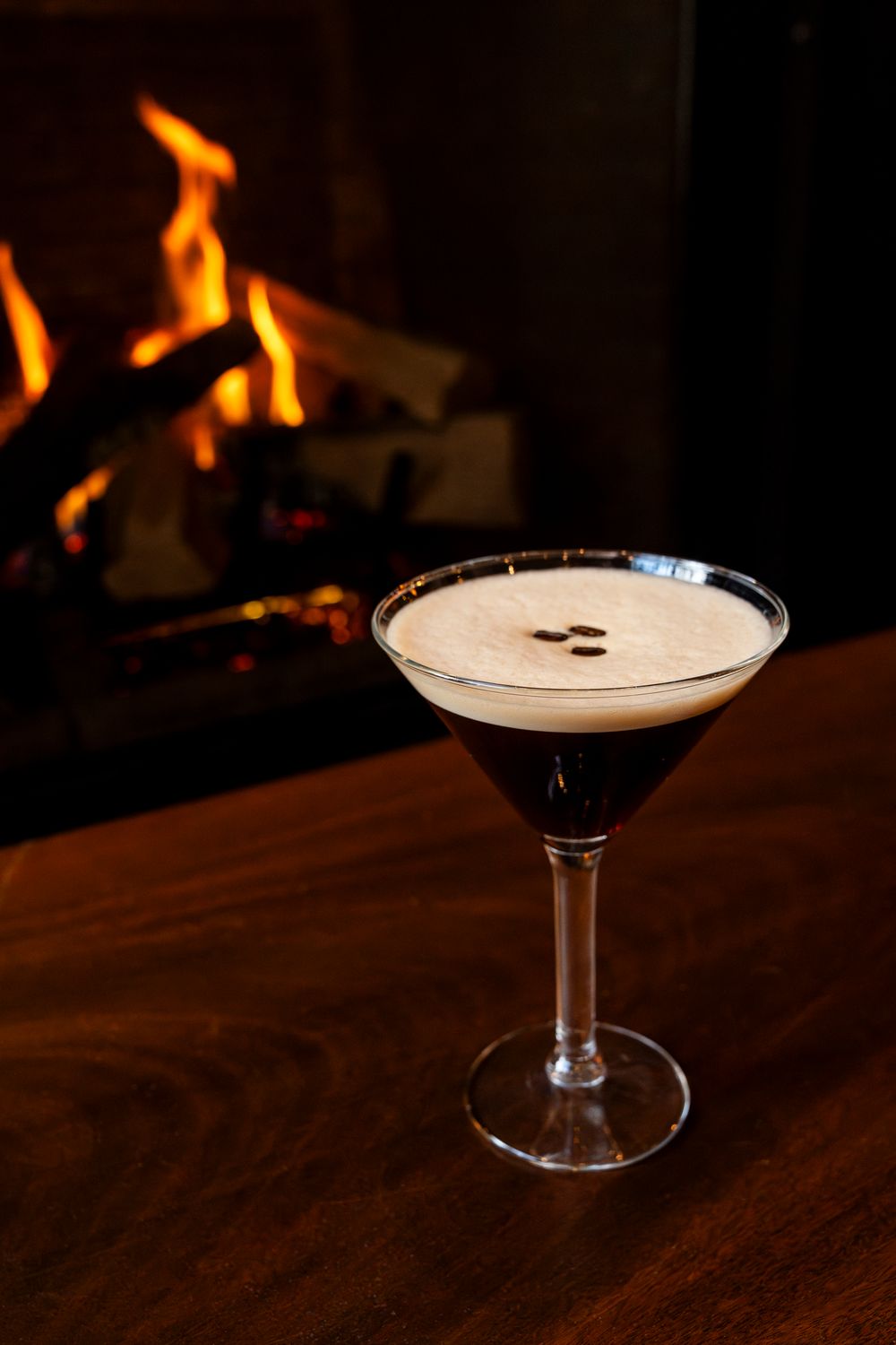 Craft cocktail with frothy top and coffee beans garnish served in front of a fireplace.