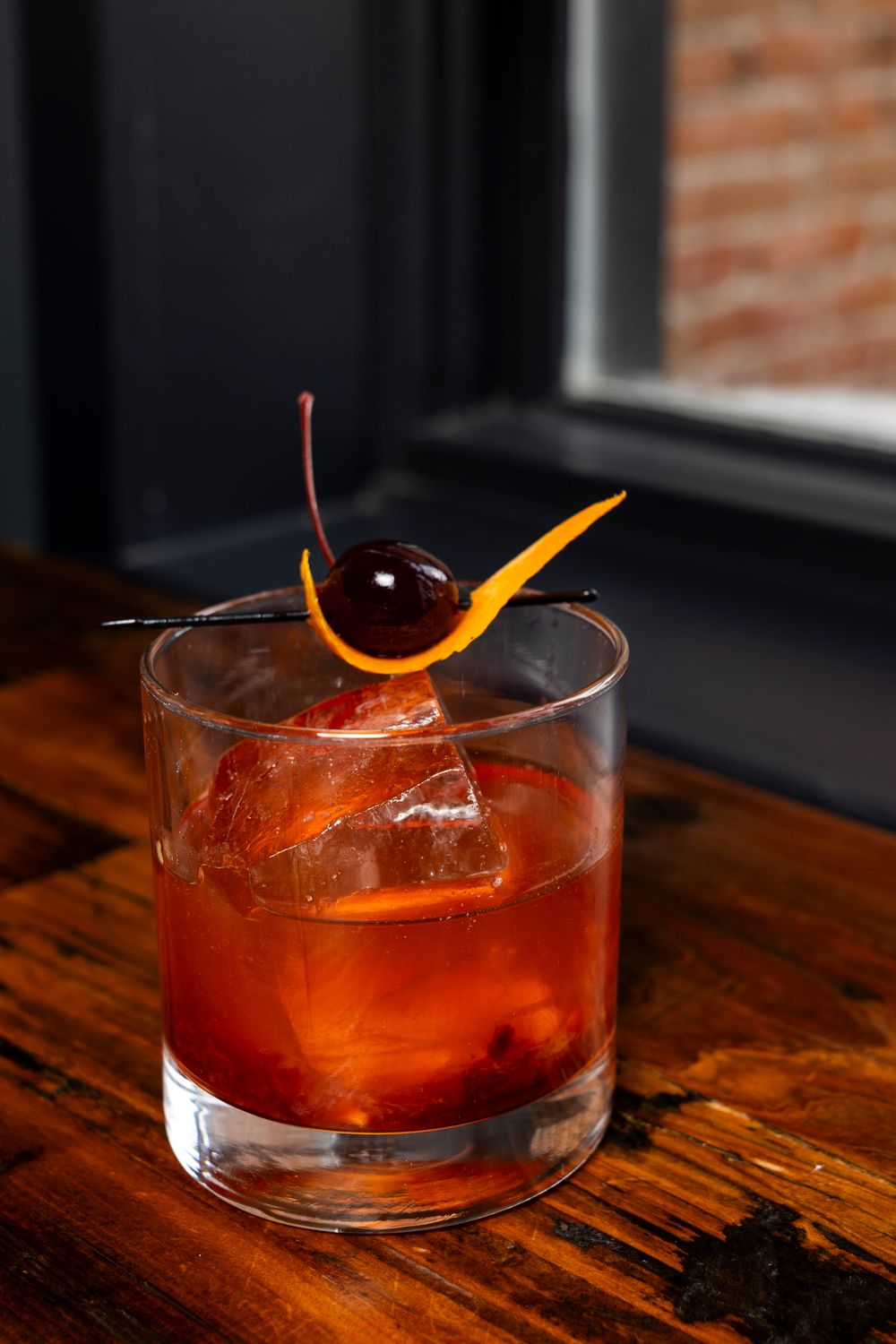 Old fashioned cocktail garnished with orange peel and cherry on wooden bar top.