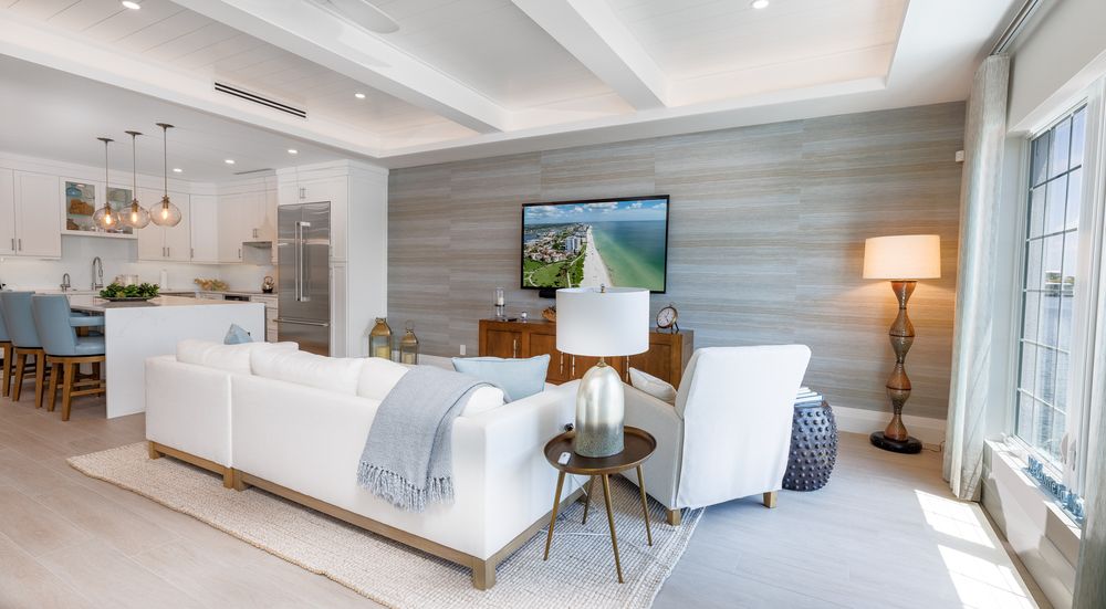Bright open-concept living room with white sofas, wood accent wall, flat-screen TV, and modern kitchen — luxury South Florida condo