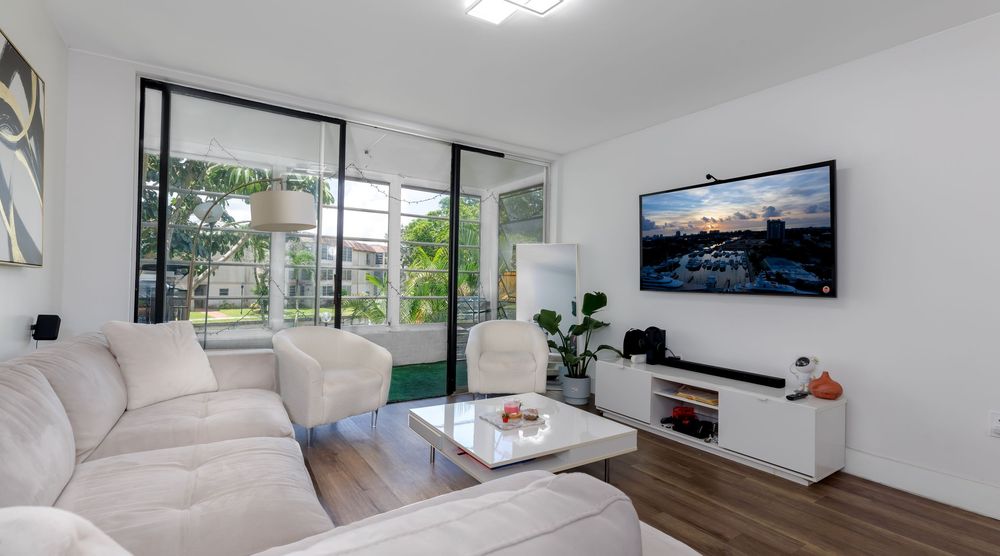 Modern living room with large windows, stylish sectional sofa, flat-screen TV, and bright natural light — perfect South Florida condo interi