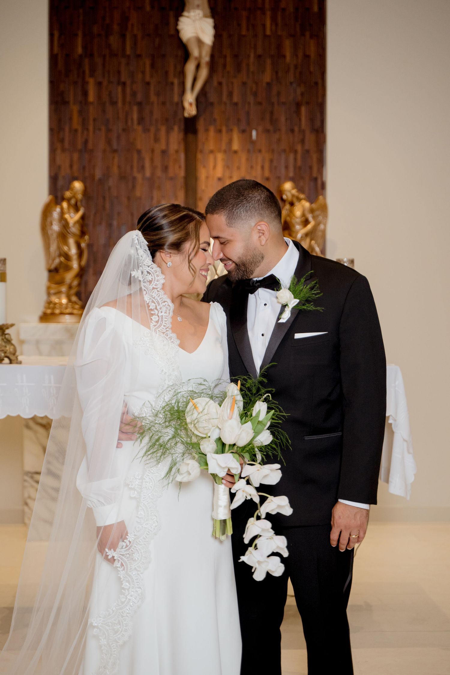 A bride in a lace gown holds a cascading white bouquet while sharing an intimate moment with her groom in a black tuxedo.
