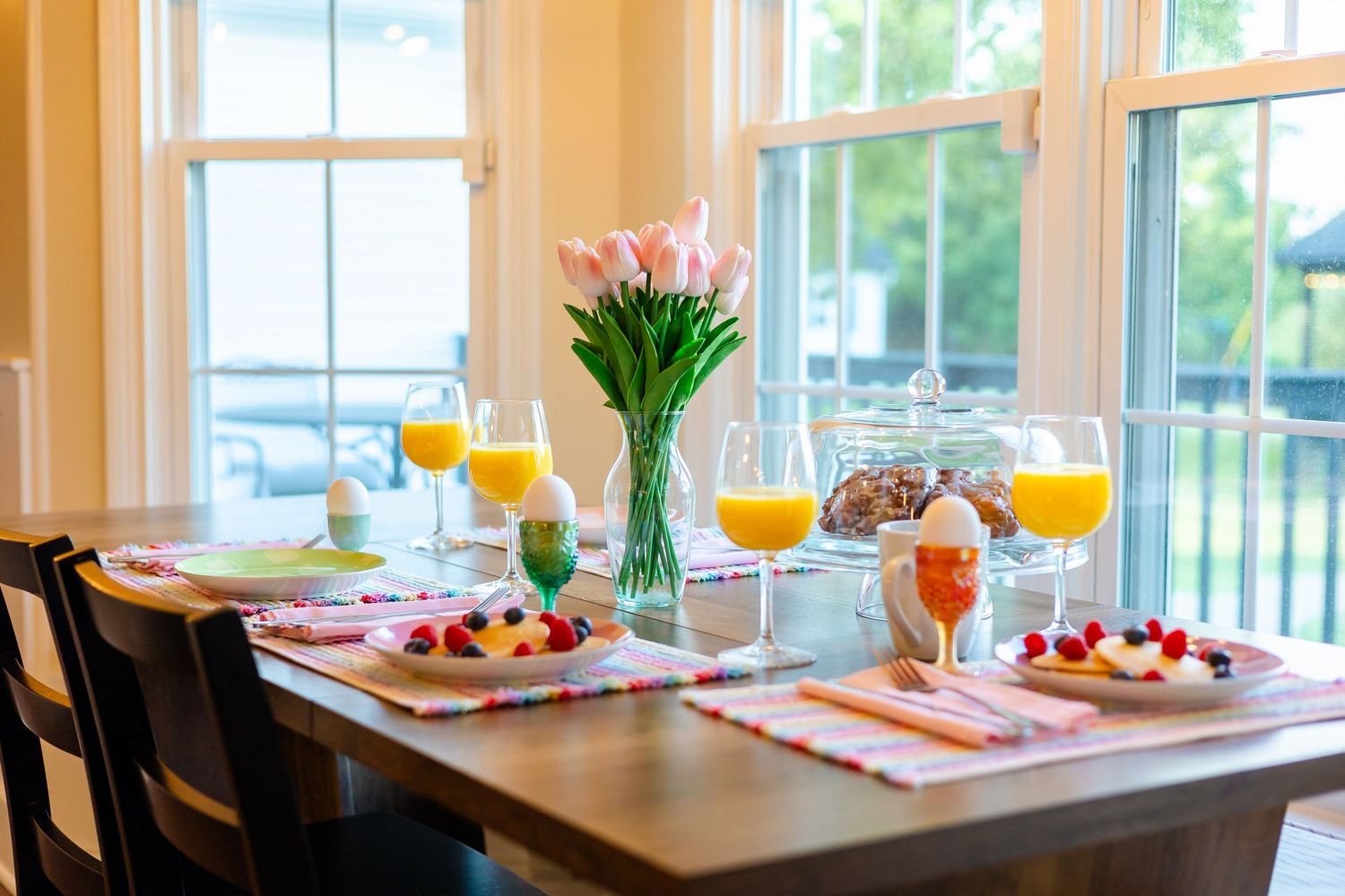 A romantic breakfast table setting with fresh tulips, orange juice, and pastries by sunny windows overlooking a garden.