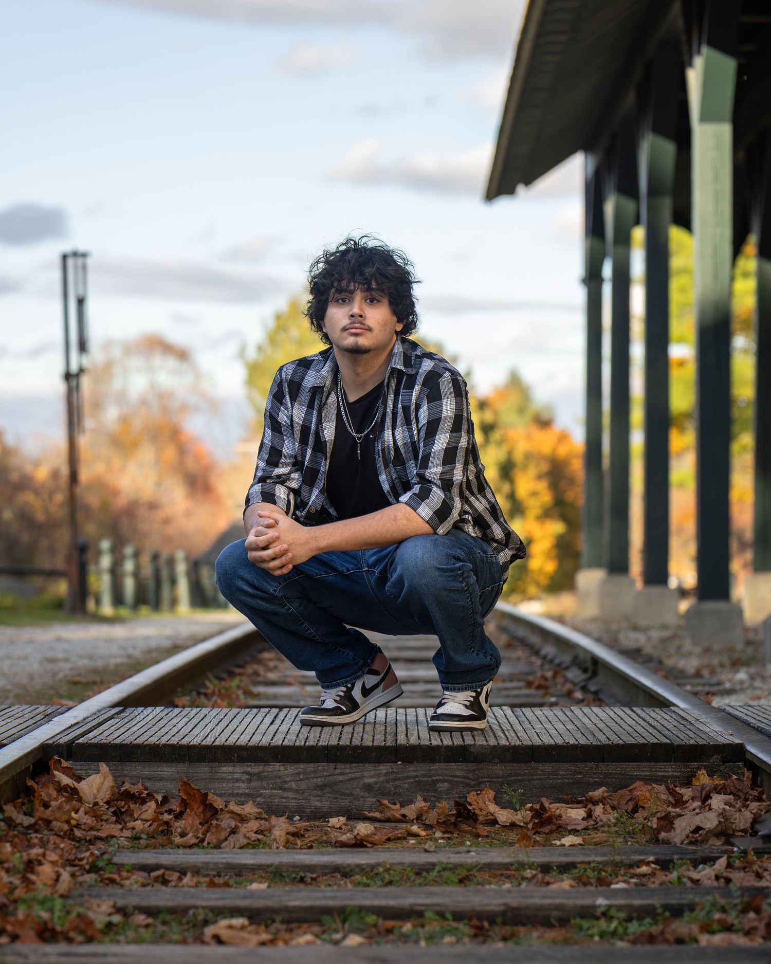 Young man in plaid shirt crouching on railroad tracks surrounded by autumn foliage.