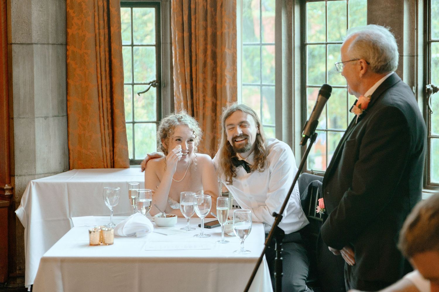 Guests gather for a formal dinner event in an elegant dining room with large windows and white tablecloths.