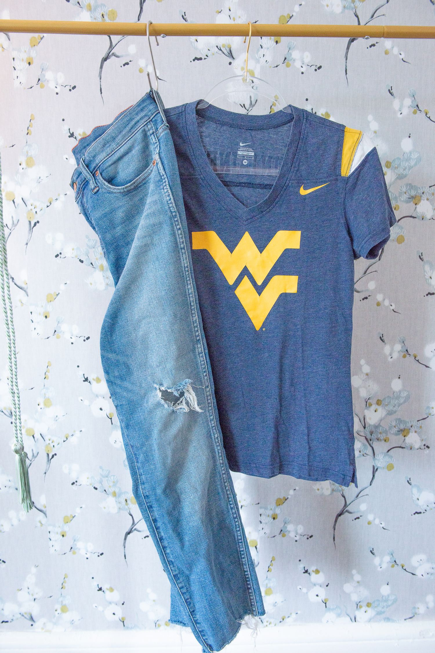 West Virginia University t-shirt with yellow WV logo paired with distressed blue jeans against floral wallpaper.