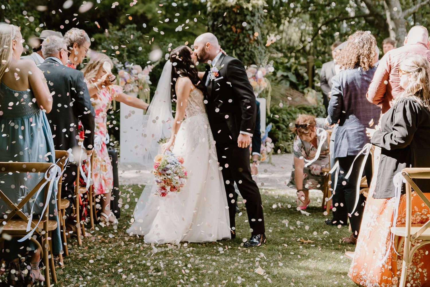 Guests shower confetti on a couple walking down an outdoor wedding aisle decorated with scattered flower petals.
