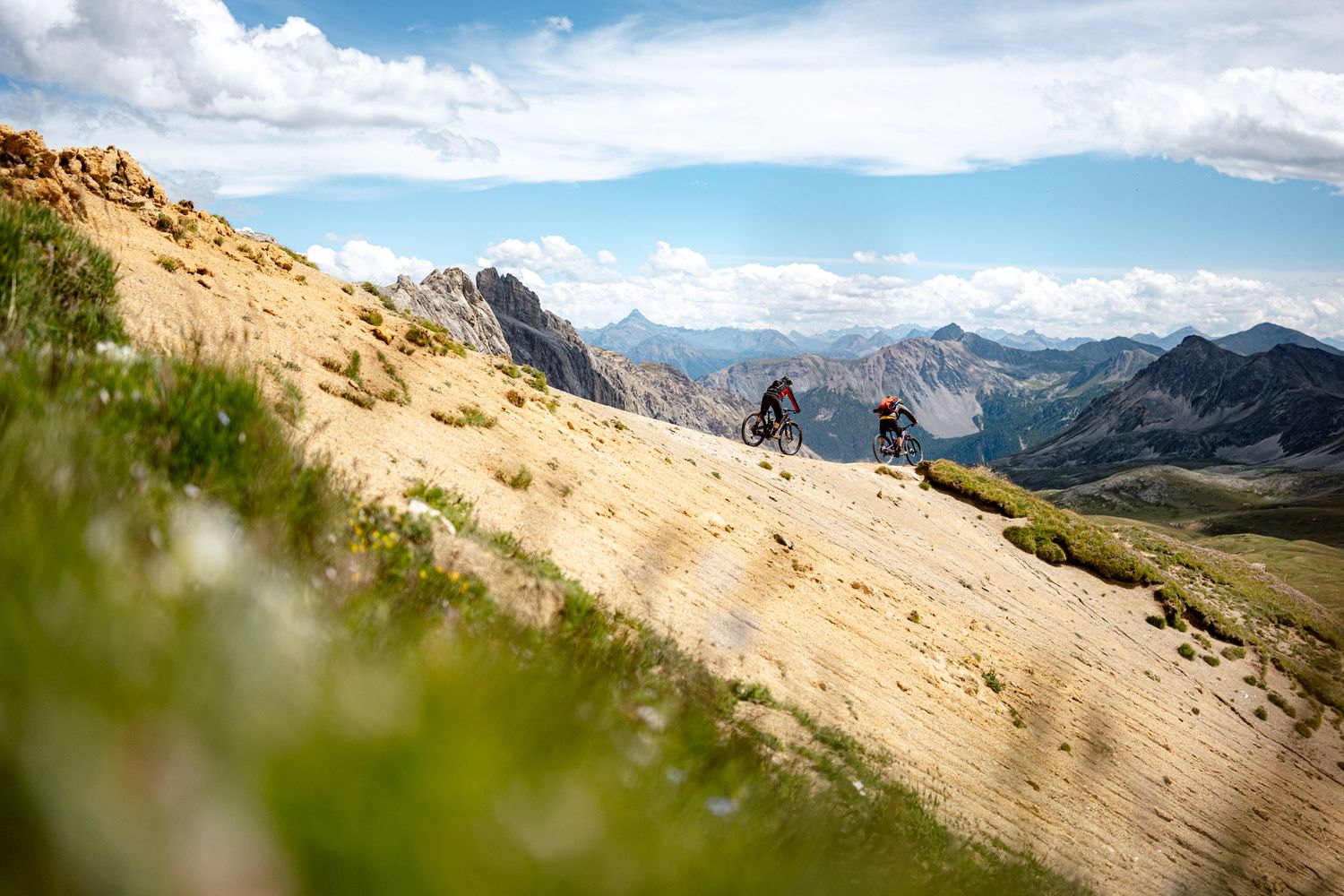 Mountain bikers traverse a scenic alpine trail on a sunny day with dramatic mountain peaks and valleys in the background.