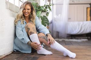Woman in denim shirt and white socks smiling on wooden floor with indoor plants, personal branding portrait in natural light.