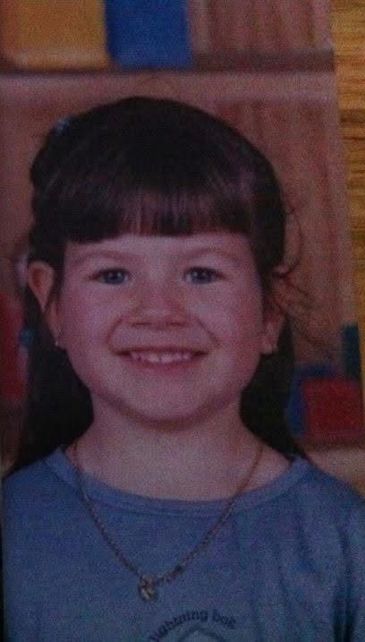 A young child in a blue shirt wearing a necklace and bangs smiles warmly at the camera.