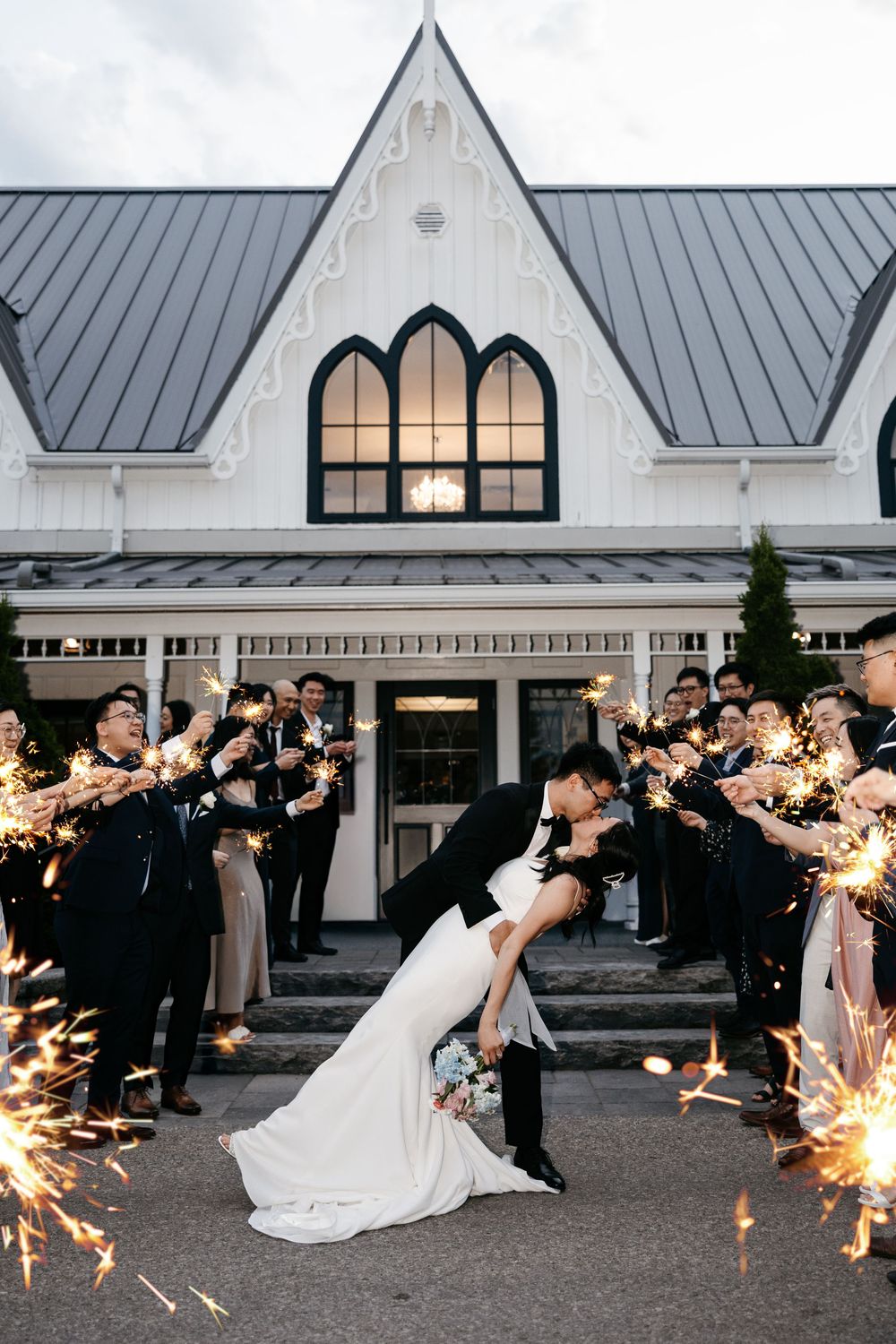 Wedding couple share romantic dip kiss outdoors in front of Gothic church while guests hold sparklers at night.