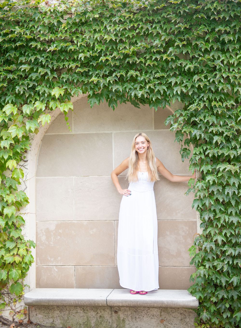 A person in a white maxi dress stands in a stone archway covered with lush green ivy vines.