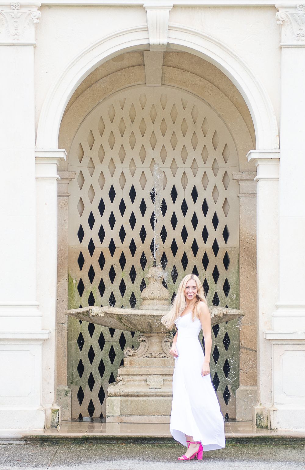 A person in a white dress poses playfully in front of a decorative stone archway with latticed window details.