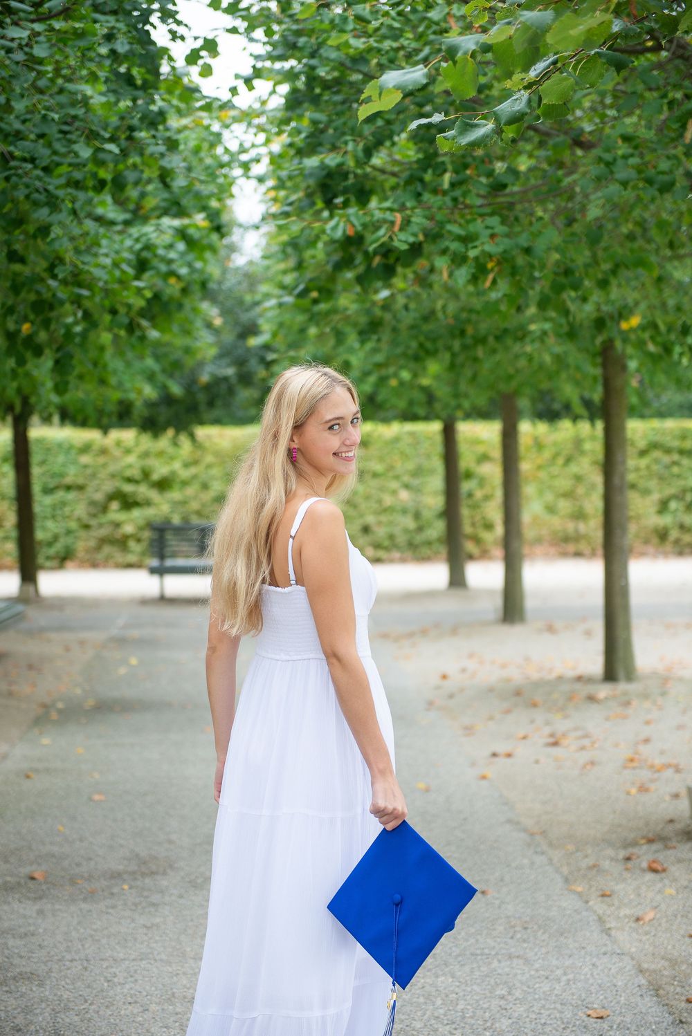 A person in a flowing white summer dress carries a bright blue clutch while walking along a tree-lined path in a park.