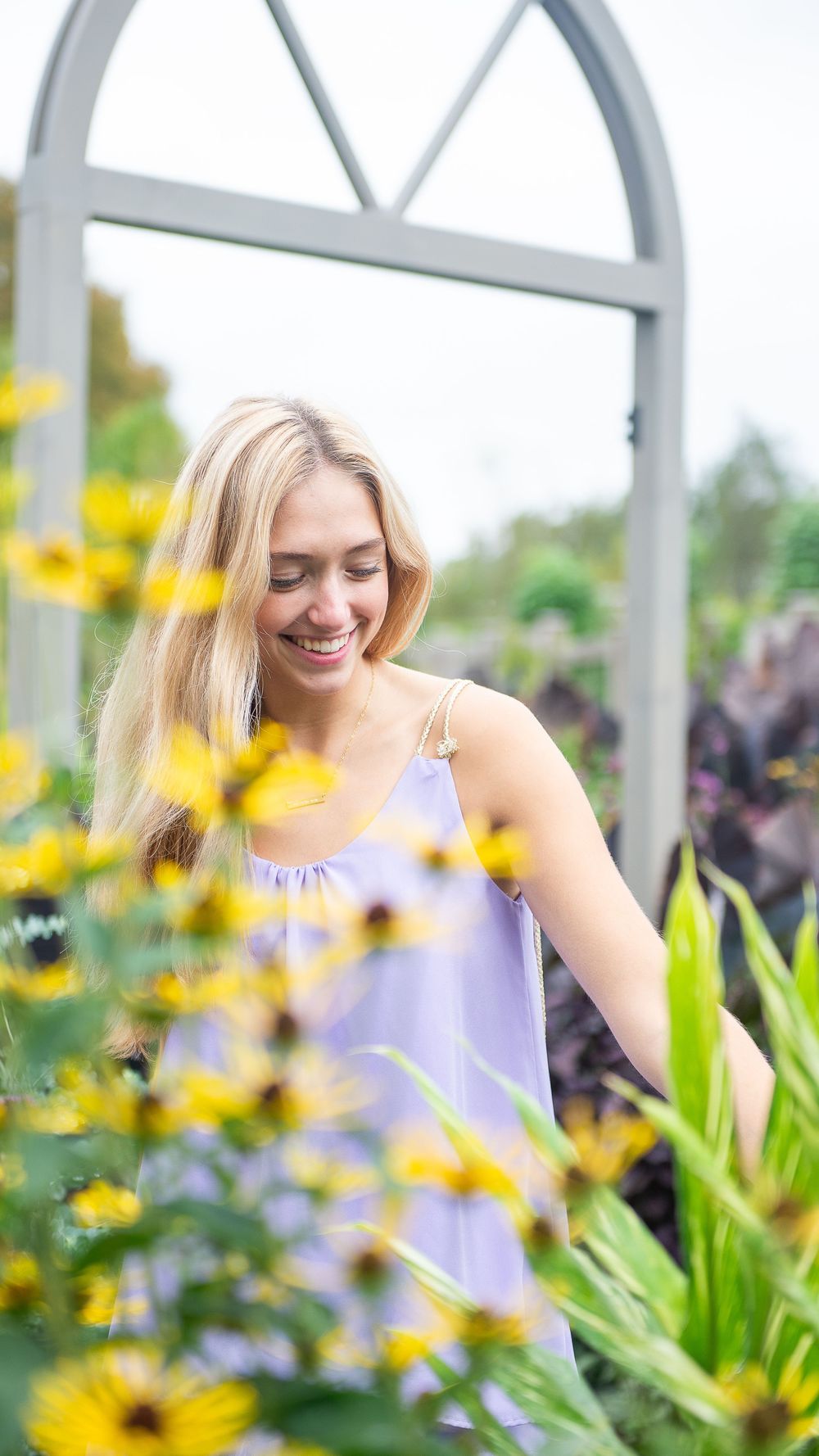 A person in a lavender dress smiles while standing in a garden with yellow flowers and a white archway.