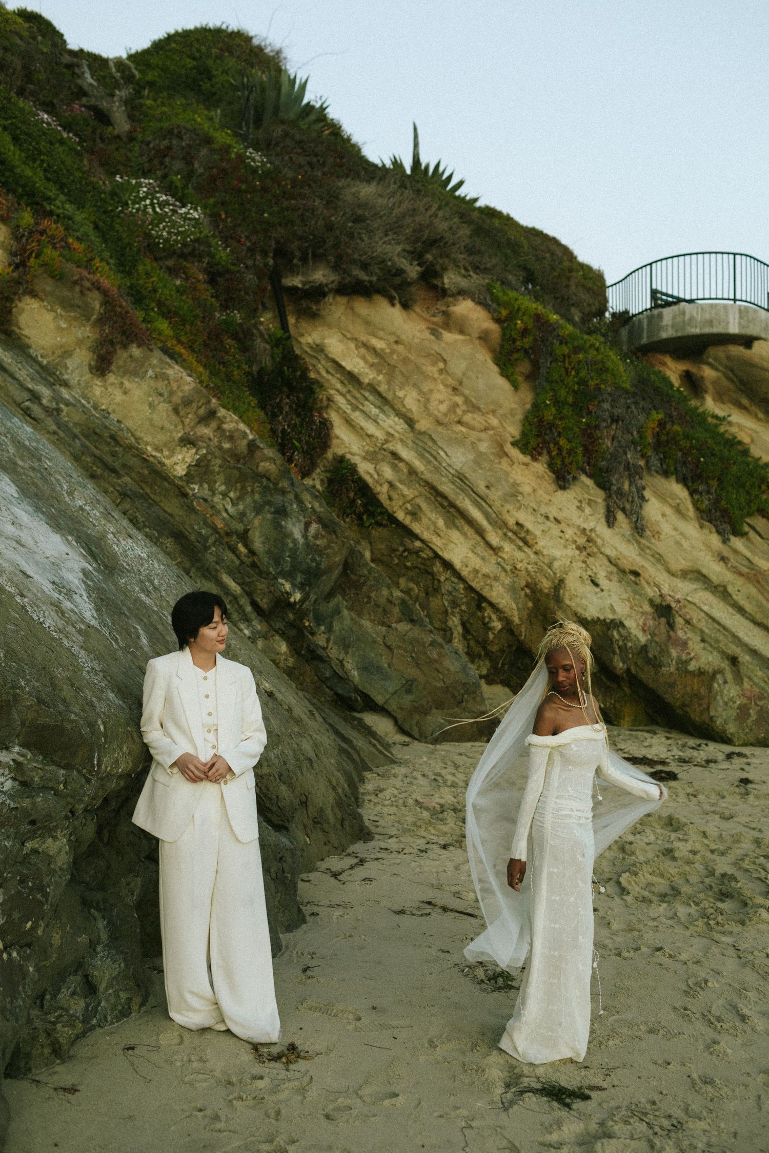 One bride leans on a rock cliff looking at their wife twirling in their veil