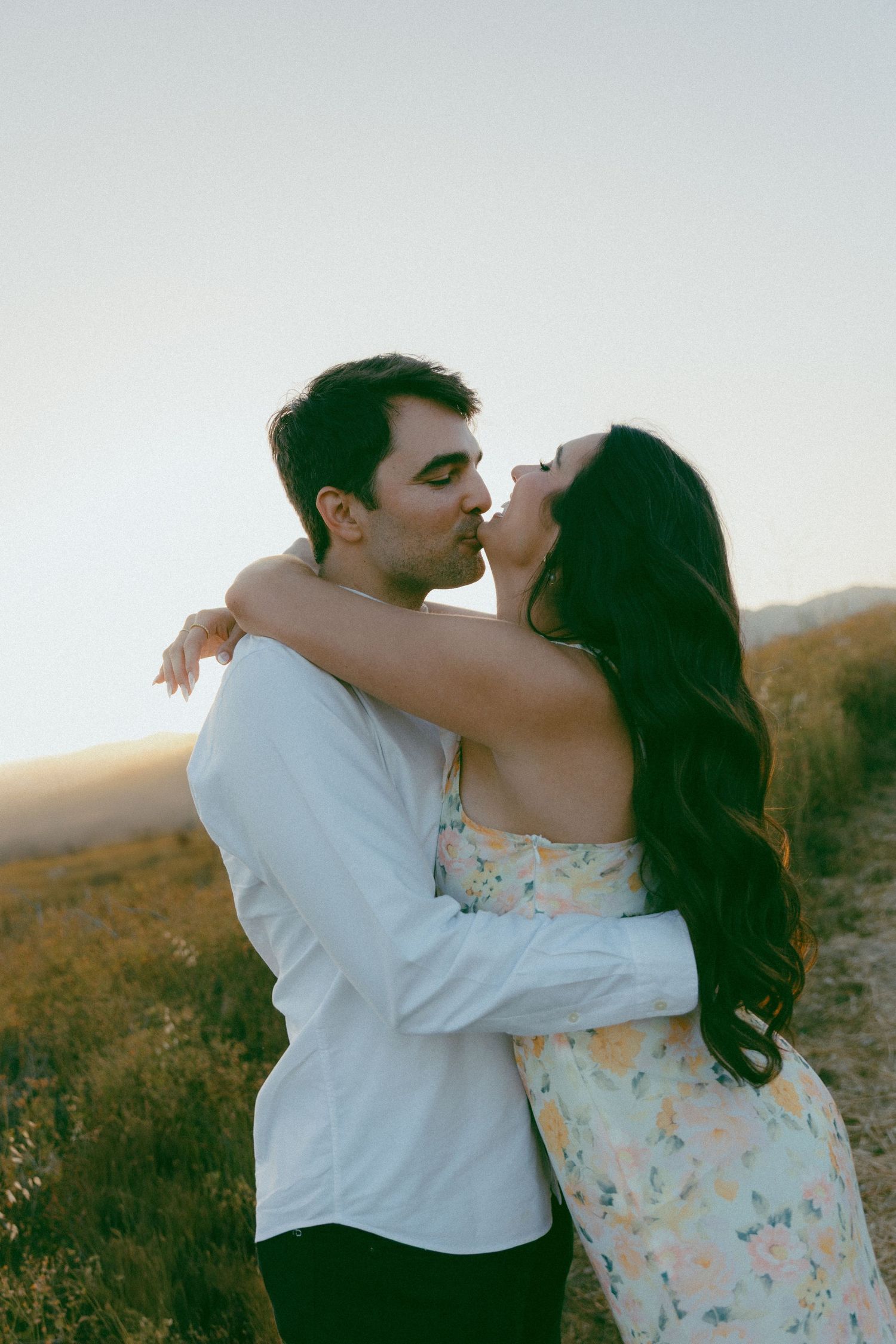 A couple embraces and kisses in a rural field at sunset wearing white and floral outfits.