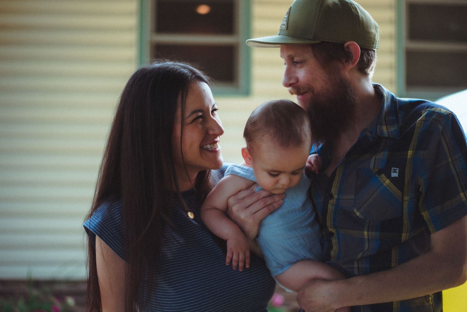 A smiling young family portrait with a mother and father holding their baby, who is looking down. Whidbey island
