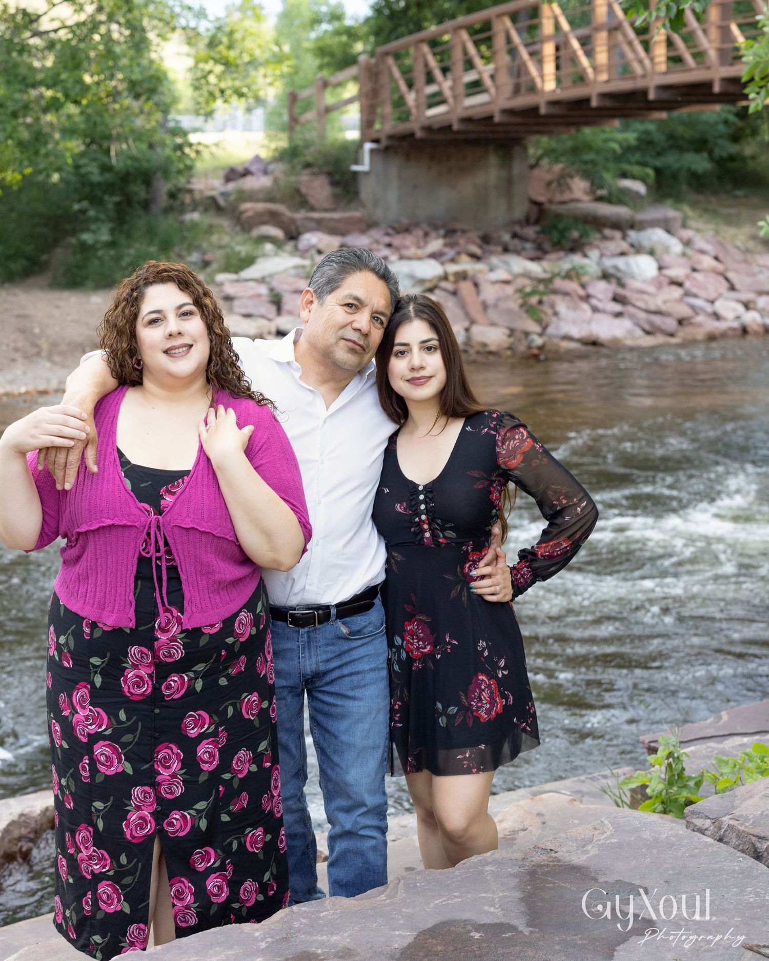 Three people standing by rushing river waters with a wooden bridge visible in the background.
