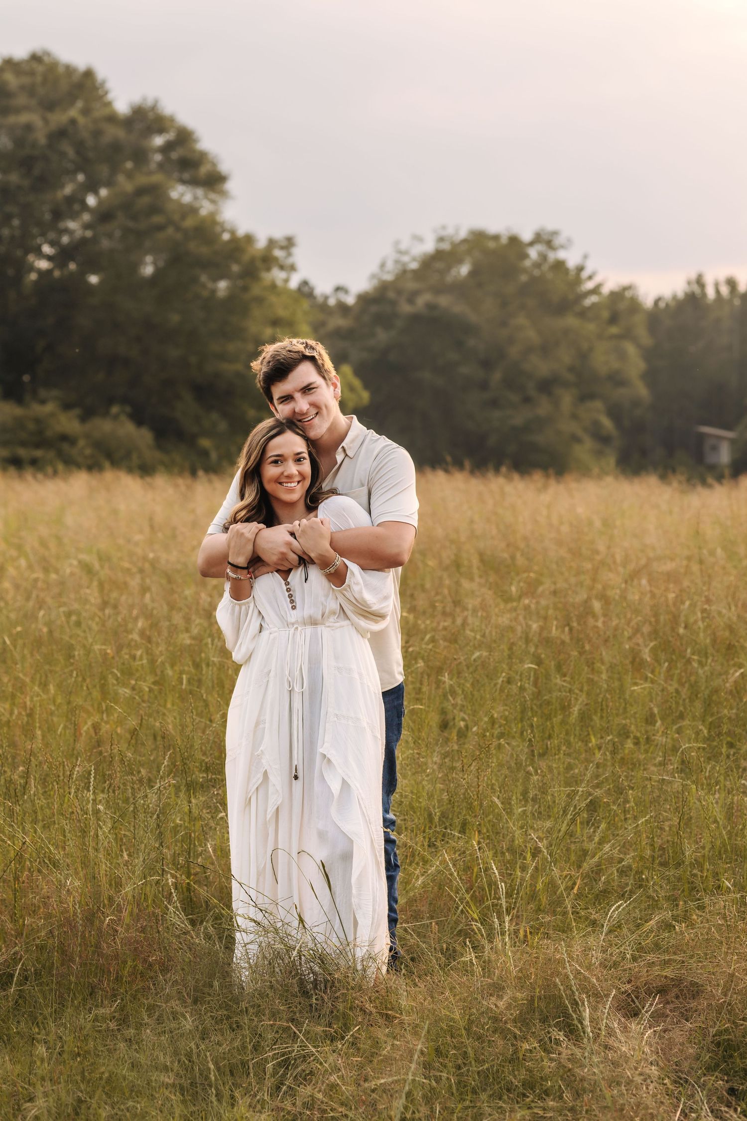 A couple embraces in a golden field at sunset with trees in the background during a romantic photoshoot.