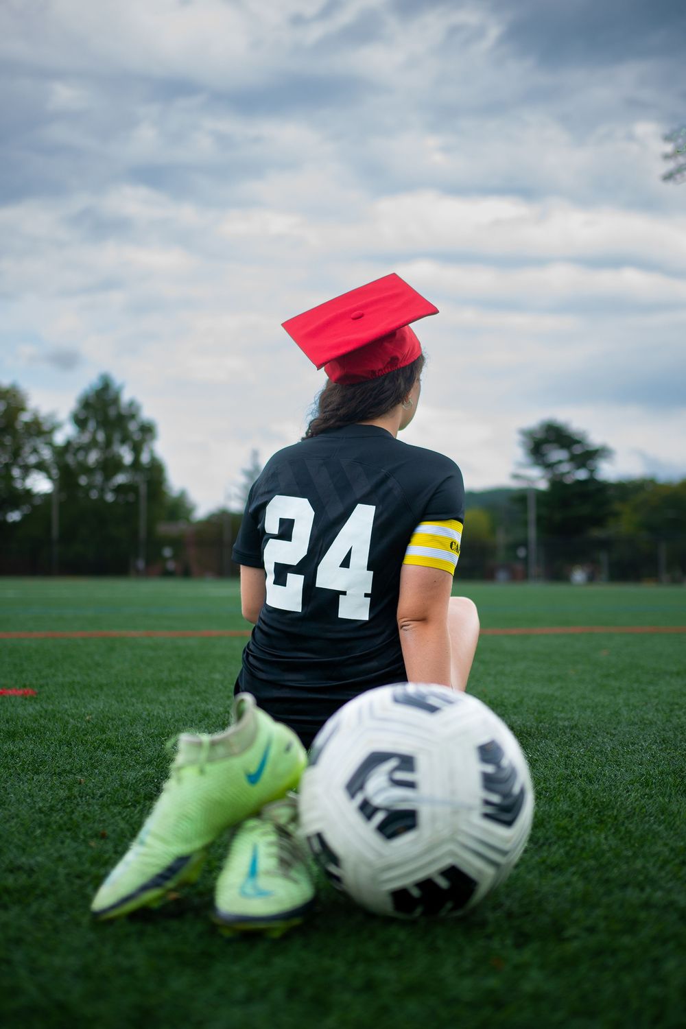 Soccer player sits on field with soccer ball and pink hat while wearing number 24 jersey under cloudy sky.