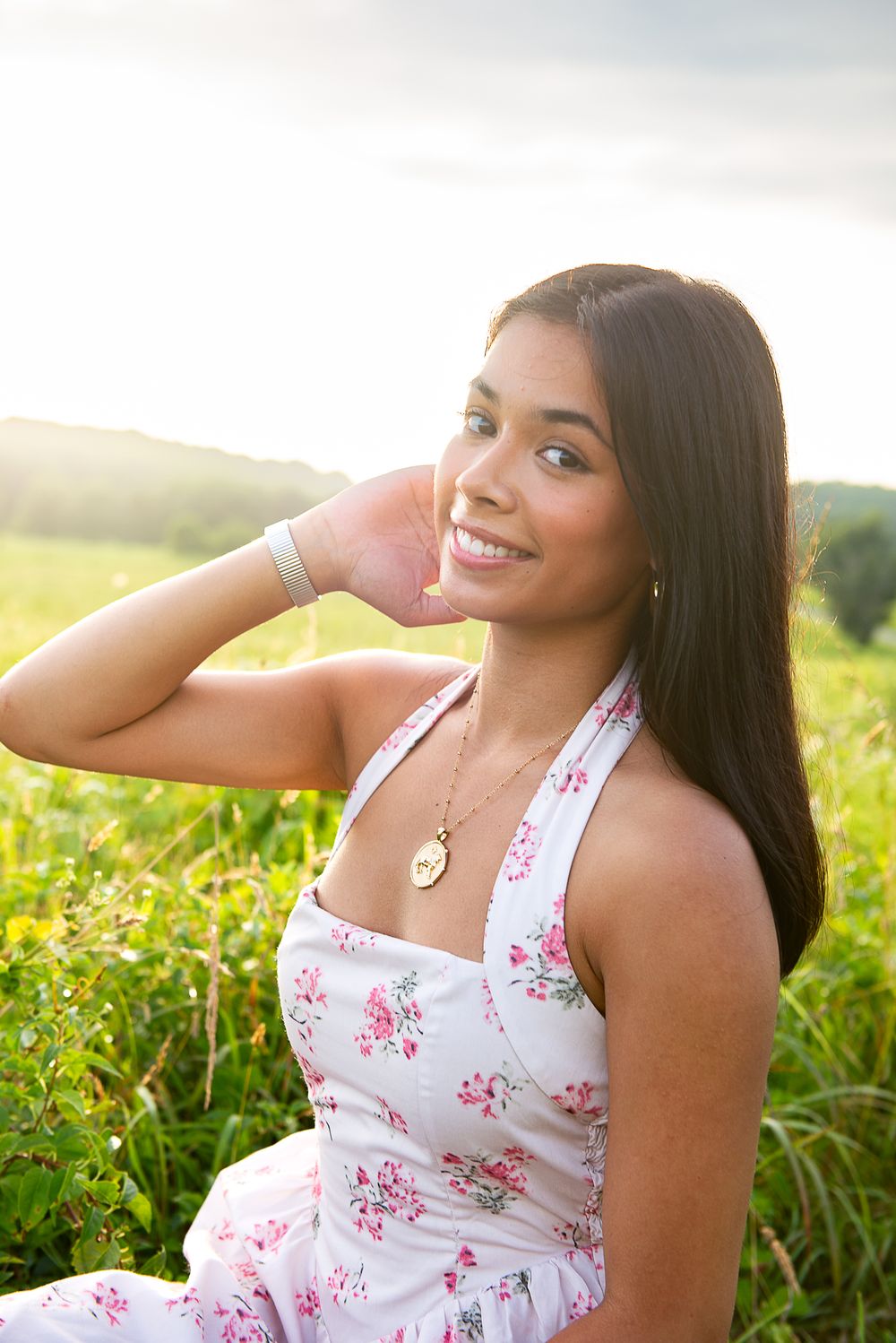 A smiling woman in a floral white summer dress sits in a sunlit meadow during golden hour.