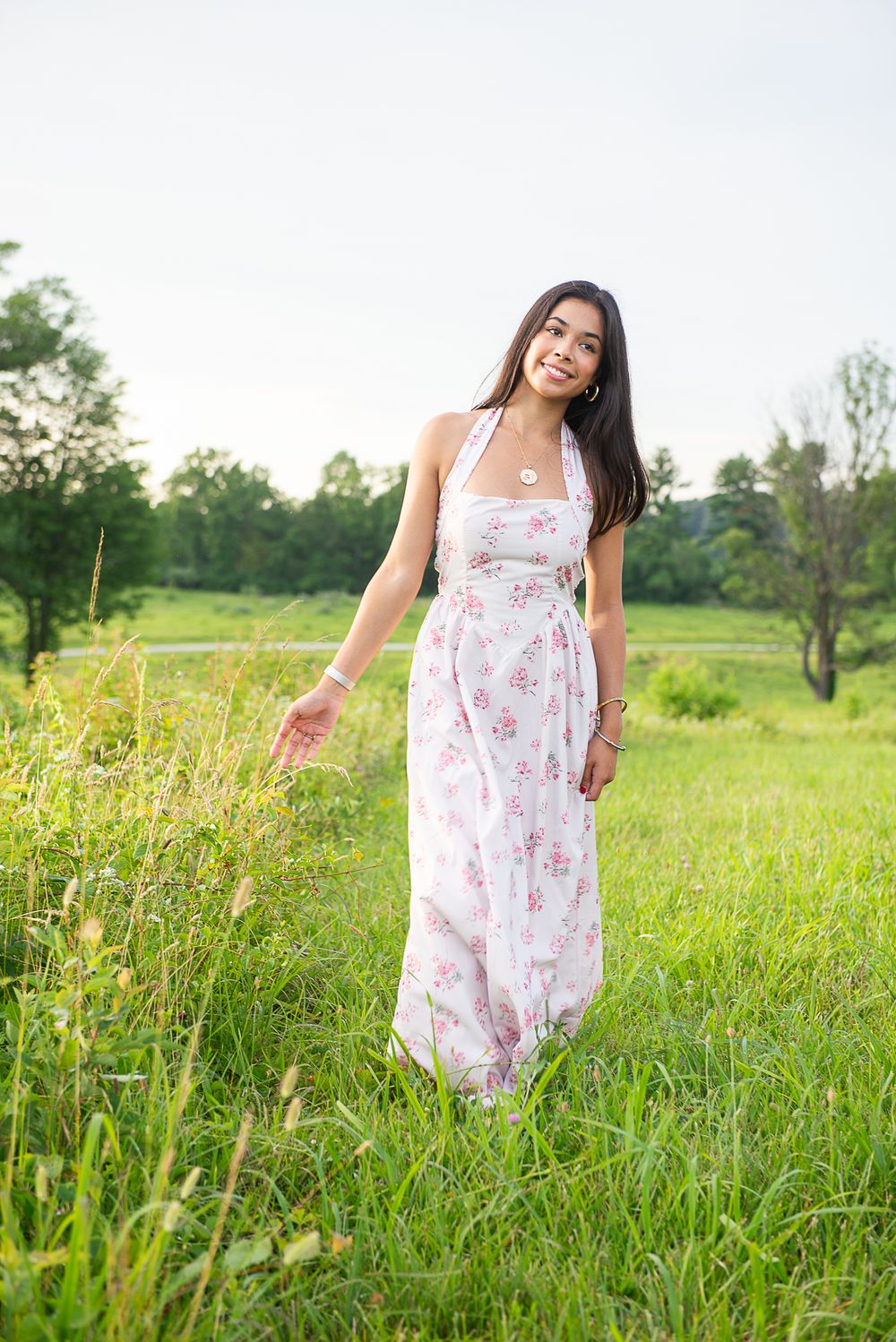A person in a floral summer dress walks through a sunny meadow field with tall grass and wildflowers.